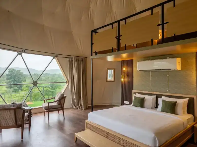 Interior of a luxury glamping dome at dusk, featuring a plush king-size bed with high-end linens, soft warm ambient lighting, and large clear panoramic windows looking out into a dense, vibrant green jungle.